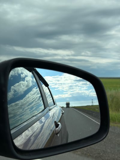 The image shows a side-view mirror of a car, reflecting a vast sky filled with dramatic, layered clouds. The mirror also captures a portion of the car's body, a black door handle, and the side window. The reflection in the mirror shows a road extending into the distance, with a green field on the right side and a power line tower. A truck is visible on the road, heading away from the viewer. The sky in the reflection is a bright blue with scattered white clouds, contrasting with the darker, more overcast sky visible in the upper portion of the main image. The overall impression is of a journey through a wide-open, possibly rural landscape. The location is specified as Kidryasovo, Russia.