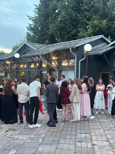 A group of young people, predominantly female, are gathered outdoors in a vertical orientation. They appear to be dressed for a formal occasion, with several individuals wearing dresses. Some are holding red ribbons. The scene is set on a cobblestone pathway next to a building with a dark facade and large windows. String lights are visible along the building's eaves. Trees with lush green foliage are in the background. The lighting suggests it is daytime, possibly late afternoon or early evening. The people's expressions are varied, with some smiling and others looking more serious. There are no visible text elements in the image.