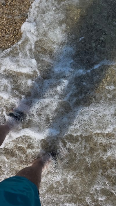 An overhead shot captures a person wading in the shallow, clear waters of a beach in Ασπροβάλτα, Greece. The lower torso and legs of an individual are visible, clad in blue swimming trunks. Dark-colored sandals or water shoes are worn on both feet, partially submerged in the foamy water. The water is highly agitated with extensive white foam and bubbles, indicating small waves breaking on the shore. Its transparency allows for clear visibility of the seabed. The visible shoreline on the left consists of light brown pebbles and gravel. Beneath the water, the seabed varies, showing a mix of lighter stones near the person's feet and darker, possibly deeper or rockier, patches further into the frame. Bright sunlight illuminates the scene, creating strong highlights and shadows on the water's surface.