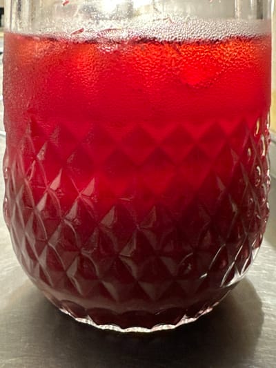The image shows a close-up of a glass filled with a red liquid, likely juice or a cocktail. Condensation is visible on the outside of the glass. The glass has a textured, diamond-patterned surface. A shadow is cast on the surface the glass is resting on. There is no indication this image was taken in Новоглаголево, Russia.