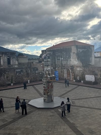 An outdoor plaza features a tall, abstract sculpture composed of small, square compartments. People stroll around the sculpture on a paved square with diagonal lines. Adjacent to the square, a weathered building stands with a red-tiled roof. A mountain looms in the background, partially obscured by the building. The sky overhead is filled with a mix of clouds, with patches of blue visible.