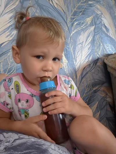 The image shows a close-up of a young child, likely a toddler, lying down and drinking from a bottle. The child has blonde hair pulled into a small bun and is wearing a white and pink onesie with a cartoon rabbit print. Their eyes are open and looking towards the viewer. The child is holding the bottle with both hands, and the bottle contains a dark liquid.
The child is lying on a bed with a blue and white patterned duvet that features a feather or leaf design with sparkly gold accents. The lighting suggests it is daytime, possibly indoors. There is no visible text in the image. The setting appears to be a bedroom. The child's expression is neutral, and the overall mood of the image is calm and domestic. The location context provided is Sokur, Russia.