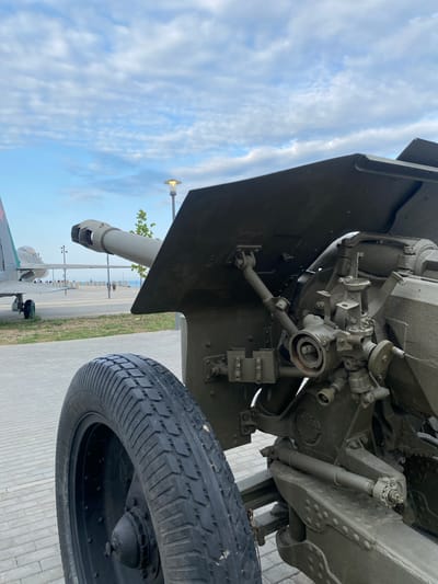 The image shows a close-up of a military cannon's wheel and part of its mechanism, with an airplane in the background. The cannon appears to be olive-drab green, as is the airplane. The scene is outdoors on a paved area, likely a promenade, with a body of water visible in the distance. The sky is partly cloudy, with a lamppost visible in the background. Several people are walking in the distance, appearing as small figures on the promenade. The location is identified as Arablyar, Russia.