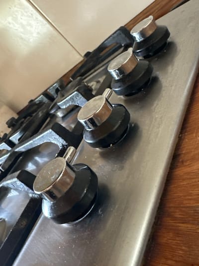 The image shows a close-up, angled view of the control knobs of a gas stove. The stove appears to be stainless steel with black knobs, each topped with a silver-colored metal cap. The stove grates, also black, are visible in the background. The surface of the stove and the knobs show some signs of use, with minor smudges and debris. To the right of the stove, a wooden surface is visible, possibly a countertop or table. The background behind the stove is a light-colored tiled wall, with the tiles appearing to be a pale beige or cream color. There are no people or animals present in the image. The lighting suggests an indoor setting, likely a kitchen. There are no specific landmarks or indicators to place this scene in the city of Trieste, Italy.