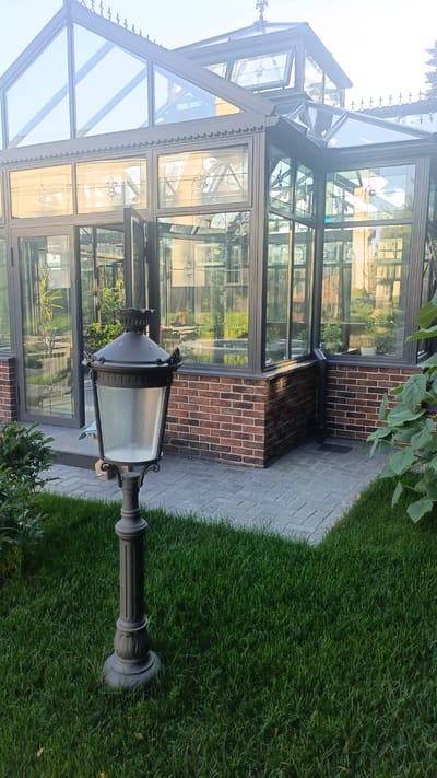 A dark grey, ornate street lamp stands in the foreground on a perfectly manicured lawn. Behind the lamp, a modern greenhouse with a dark grey frame and a brick base is visible. The greenhouse has large glass panels, some of which are open, allowing a view of the lush greenery and plants inside. The roof of the greenhouse is also made of glass, with decorative finials along the top edge. The ground around the greenhouse is paved with grey interlocking bricks.
The scene appears to be set in the late afternoon or early evening, as the light is soft and casts gentle reflections on the greenhouse glass. The weather seems pleasant, with a clear sky visible through the glass roof. The overall impression is one of a well-maintained garden with a touch of elegance. The location is suggested to be Nikolino, Russia. There is no visible text in the image.