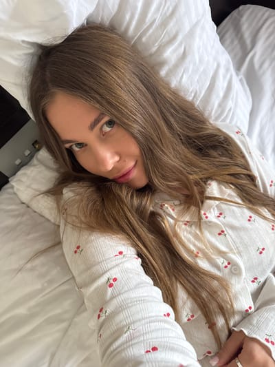 A woman is lying on a white bed. She has long brown hair and is wearing a white shirt with cherry patterns. The location cannot be determined as Sosnovy, Russia, from the provided image.