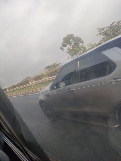 The scene, located in Murera ward, Kenya, depicts a dark-colored SUV, consistent with a Land Rover Discovery 5 model, traveling on an asphalt road. The SUV exhibits tinted windows on its passenger side. Adjacent to the road, a concrete barrier with a metal railing extends along the thoroughfare, separating it from an upward-sloping embankment. This embankment features sparse green vegetation and several trees. The background is dominated by an overcast, light gray sky. No individuals are visible. The image is captured from the interior of another moving vehicle, with a blurred, dark reflection occupying the immediate left foreground.