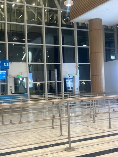 This image captures a modern indoor public space, likely an airport terminal in Addis Ababa, Ethiopia. The scene is dominated by a grid of large glass windows framed in white, extending across the left and center. Through these windows, reflections of the interior architecture and various light sources are visible, and the outside appears dark, suggesting evening or nighttime.

In the foreground and to the right, a long series of silver metal queue barriers with connecting bars is arranged on a light-colored tiled floor, which features alternating dark and light stripes. These barriers suggest an area for passenger queuing, although no people are visible.

A prominent light brown, ribbed, cylindrical fixture hangs horizontally from the white ceiling, which also features recessed lighting. On the far left, a spherical light fixture with a textured surface emits a soft glow.

Several digital display screens are visible. One blue screen in the lower left clearly shows "C5," likely a gate number, with smaller text below indicating flight details: "22:50," "ET 985," and "23:50." The "ET" likely refers to Ethiopian Airlines, supporting the given location context. Another screen, visible through the windows, reflects mirrored text that appears to be "WELCOME." The overall atmosphere is calm, orderly, and well-lit by artificial sources.