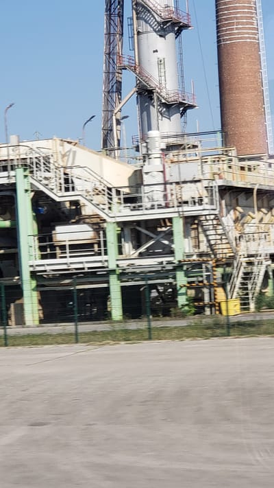 The image shows an industrial facility in Trenčianska Teplá, Slovakia. The facility features a large, metallic structure with multiple levels, walkways, and stairs, suggesting complex industrial processes. A tall, cylindrical brick chimney stands to the right of the metallic structure. The sky is clear blue, indicating daytime. The foreground consists of an asphalt surface bordered by a green fence and some vegetation. There are no people visible in the image. The overall scene depicts an operational or disused industrial site.