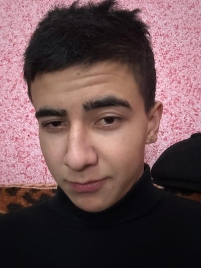 A close-up portrait features a male subject, positioned indoors in Dehqonbozor, Uzbekistan. The individual has short, dark, textured hair, thick dark eyebrows, and dark eyes with a direct, slightly downcast gaze. His lips form a subtle smirk. He wears a black turtleneck sweater. The background consists of pink, speckled wallpaper, with a horizontal section of dark brown and black animal print fabric or wallpaper visible below the main pink pattern. A black peaked cap is partially visible behind the subject's right shoulder.