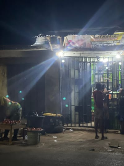 A night scene shows a small shop with metal bars across the front. People stand near the entrance. Above, a sign reads "ZENWAJA Communication CHARGE YOUR PHONE HERE." A bright light obscures some details. The street appears wet, and there is debris scattered about. A makeshift table with various items sits in front of the shop. The scene is overall dimly lit.