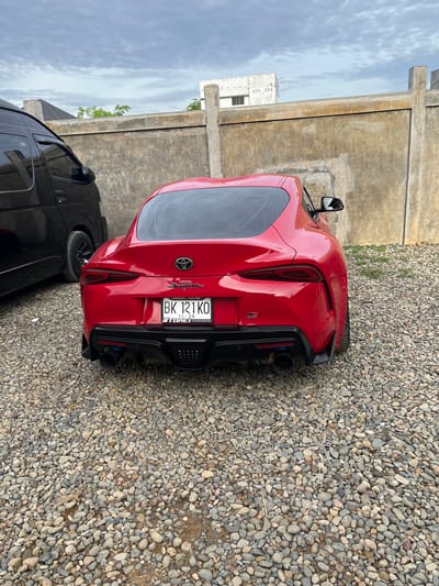 The image shows the rear of a red Toyota Supra parked on a gravel surface in Lampeuneurutgampong, Indonesia. The car has a black rear diffuser, exhaust pipes, and a license plate that reads "BK 121 KO". A black van is parked next to the Supra. Behind the vehicles is a concrete wall, and beyond that, the sky is visible.