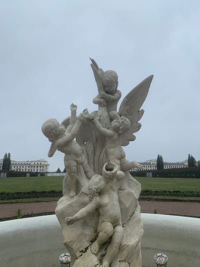 A close-up, slightly angled shot shows a white stone sculpture featuring several cherubs or putti figures, some with wings. The sculpture appears to be part of a fountain or similar decorative structure. Behind the sculpture is a grassy area, a reddish-brown pathway, and a large, cream-colored building with multiple stories and windows. The sky is overcast and gray. The scene is located in Arkhangel'skoe, Russia.