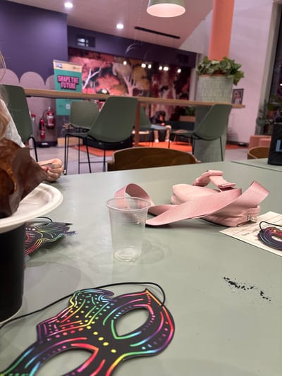 A light green table with a pink purse, a clear plastic cup, a scratch art mask, and a black and white card. The table is set in a café, with green chairs and brown tables. There is a mural behind the table. A plate and a dark object are in the foreground, partially obscuring the view. The scene is indoors.