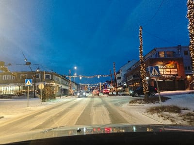 The image displays Storgata in Moelv, Norway, during low light conditions, consistent with early evening or nighttime in winter. Snow covers the ground, rooftops, and tree branches. The two-lane asphalt road exhibits visible tire tracks through a thin layer of snow. Numerous bright yellow-white decorative string lights are draped in garlands across the street and wrapped around poles or tree trunks lining the roadway.

On the left side of the street, a two-story building is visible, featuring a prominent sign that reads "Moelv Burger." Further down the street on the left, another vehicle is dimly discernible with its headlights on. A blue square pedestrian crossing sign (white triangle with a black pedestrian symbol) is erected on the left side of the road, near a partially obscured circular sign.

On the right side of the street, a multi-story building displays the illuminated sign "PASSASJEN." A dark-colored SUV is parked on the right shoulder, partially obscured by a light-wrapped pole. Another blue square pedestrian crossing sign is also visible on the right. In the distance, multiple vehicles are present, some with illuminated headlights, indicating active traffic. The sky above is a deep blue, with no discernible cloud cover. Reflections consistent with a vehicle's dashboard or windshield are visible across the bottom of the frame, suggesting the image was captured from inside a moving or stationary vehicle. No individuals are visible in the scene.