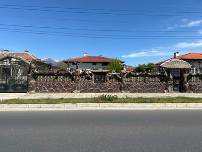 The image depicts a street scene in Gabarevo, Bulgaria. The foreground is an upside-down view of the road and sidewalk. Beyond the sidewalk is a decorative fence made of brown, rock-like material with sculpted tree-branch designs. Behind the fence are houses with red-tiled roofs. A mountain is visible in the background. Power lines cross the blue sky. There are no people visible in the image.