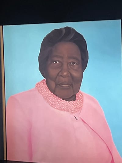 This is a portrait painting of an elderly Black woman with dark hair, wearing a pink outfit with a beaded neckline. She is depicted from the chest up against a solid light blue background. The painting is framed by a dark border. There is no discernible location in Liverpool, United Kingdom, or any specific action or interaction depicted.