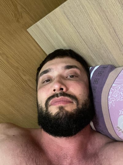 A selfie of a man with a beard and dark hair is taken from a close-up perspective, lying on his side on a bed with a wooden-patterned headboard or wall behind him. He is wearing a pillow, which has a purple and pink floral pattern, underneath his head. His skin appears flushed, particularly around his neck and chest. The lighting suggests it is indoors, possibly during the day, with natural light source from the left. The background includes the wooden texture of the headboard and a glimpse of what appears to be a wooden floor.