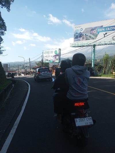 The image depicts a daytime scene on an asphalt road in a mountainous or hilly area. Two individuals are seen from behind, riding a motorcycle in the foreground. The passenger wears a light grey hooded sweatshirt with a crest containing a red symbol, text, and "COUTAS" below; both riders wear dark helmets. The motorcycle's rear is visible, showing a red LED taillight assembly and an Indonesian license plate, B 3867 CYM, with "12-2G" below the number.
The road curves slightly to the left, with a solid white line marking the left edge and a faded yellow line near the center. A concrete drainage ditch runs alongside the left road edge. Ahead, a silver SUV is observed navigating the curve. In the background, two large billboards are prominent. The closer billboard advertises "LEMBAH HIJAU - RESORT HOTEL CILOTO." The larger billboard further back displays "BBPK CILOTO" alongside imagery of modern buildings. Power lines are visible strung across the scene. The distant background shows more hills or mountains under a partly cloudy blue sky, with scattered buildings indicating a populated area. Based on the license plate format and the "Ciloto" references on the billboards, the location is identified as Ciloto, West Java, Indonesia.
