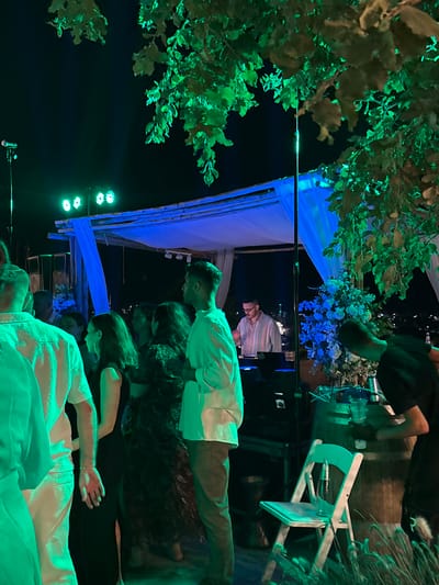 This image shows a nighttime outdoor event with several people gathered. The scene is illuminated by green and blue lights, with some of the lighting cast upon the lush green leaves of a tree in the foreground. In the background, there is a structure draped with blue fabric, possibly a tent or canopy, and a white folding chair. Several people are visible, some standing and others appearing to be in conversation or observation. One person in the foreground is wearing a white, textured shirt. Another person is wearing a white long-sleeved shirt. There is a floral arrangement, predominantly blue flowers, displayed on a table. The overall atmosphere suggests a celebratory or social gathering. The image is oriented vertically.