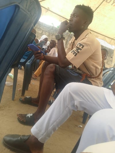 The image depicts an outdoor gathering in the city of Ijoko, Nigeria, under a light-colored canopy. The ground is exposed earth. In the mid-ground, a dark-skinned male with short hair sits on a dark blue plastic chair. He wears a light tan short-sleeved shirt with "EME" in black on the right sleeve, dark shorts, and dark slide sandals with a white logo. A dark smartwatch is on his left wrist. He holds a blue smartphone in his left hand, connected by a dark cable, and his right hand is raised to his mouth. A braided, rope-like accessory with a tassel hangs from his neck. His right knee exhibits a noticeable scar.
In the immediate foreground, the lower legs of another seated individual are visible, clad in white trousers and dark leather slip-on shoes with buckles. To the left, a portion of another dark blue plastic chair is visible. In the background, several individuals are seated on similar chairs. A young female wearing a light head covering and top looks towards the viewer. Another person with a head covering is partially obscured. Behind them, a light-colored wall structure is visible. Small debris, including a green plastic bottle, lies on the ground.