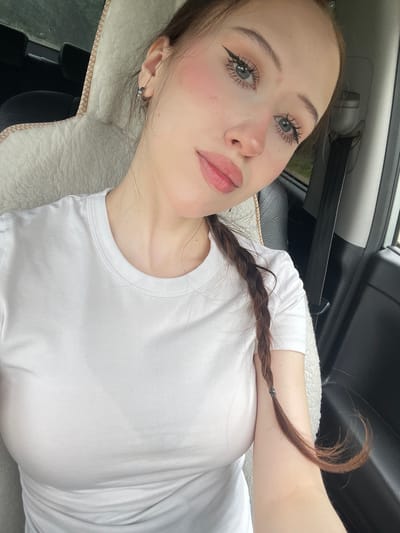 In Gavrilovka, Russia, a young woman with fair skin, light blue eyes, and dark brown hair braided into a pigtail sits in the passenger seat of a vehicle. She is wearing a plain white t-shirt. Her makeup includes winged eyeliner, mascara, and blush. The car's interior is visible, with a fuzzy white seat cover behind her and a black seat and seatbelt to her right. The focus is on her face and upper body as she takes a selfie.