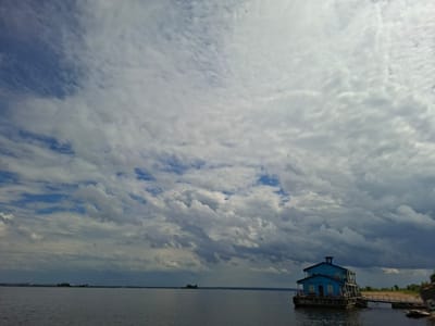 A wide shot captures a body of water under a dramatic sky filled with heavy, textured clouds. The clouds range from bright white to dark grey, creating a sense of impending weather. In the foreground, on the right side of the image, sits a weathered, blue two-story building that appears to be a houseboat or floating structure. It is docked by a ramp leading to a sandy shore with sparse vegetation. The building has visible windows, doors, and a chimney. Across the water, in the distance, is a faint outline of land, possibly the city of Verkhny Uslon, Russia, with a small, tree-covered island visible in the middle ground. The water is dark and calm, reflecting the moody sky. The overall impression is one of a slightly desolate, yet picturesque, scene on the water.