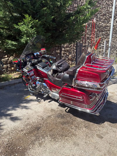 A parked, cherry red Honda Gold Wing motorcycle with extensive chrome detailing and a large windshield is the primary subject. The motorcycle is positioned on an asphalt surface, which appears damp in places, with scattered small stones. Behind the motorcycle, a stone wall forms a textured backdrop, with a dense green evergreen tree to the left. A dark-colored bag is placed on the rear seat of the motorcycle. To the right of the motorcycle, a metal fence is visible, and beyond it, red poles with small bottles and a red flag on a thin pole are present. The scene is illuminated by bright sunlight, casting distinct shadows on the ground. The motorcycle is in Sotonići, Montenegro.