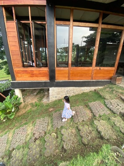 The image shows a young child in a light-colored dress with a floral pattern walking along a paved pathway next to a building. The building has a wooden facade with large windows. The pathway is made of gray hexagonal pavers and flower-shaped stones, with grass growing in between the stones. The location is in Tamiajeng, Indonesia.