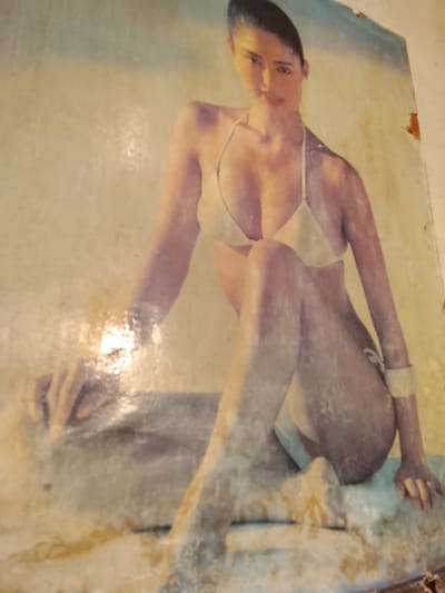 The image is a faded, possibly vintage, photograph or painting of a woman in a light-colored bikini. She is seated and appears to be posing. The background is a light, textured color, possibly a wall or a backdrop. There are areas of damage and discoloration on the surface of the image, with peeling and staining visible, particularly on the edges and in the lower portion. The lighting is soft, creating highlights and shadows on the woman's skin and the surface she is sitting on. The image does not contain any identifiable landmarks or elements that would place it specifically in Namangan City, Uzbekistan.