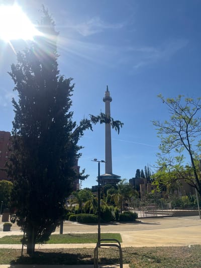 The image depicts the Torre de Collserola, a telecommunications tower designed by Norman Foster, situated in the Collserola mountain range near Sant Just Desvern, Spain. The tower, a slender concrete structure topped with an antenna, stands tall against a clear blue sky. In the foreground, a large, dark green evergreen tree with a dense canopy partially obscures the view of the tower. Sunlight streams intensely from the upper left corner, casting lens flares across the image and highlighting the foliage of the tree.
The tower's base features a circular structure with a glass facade. Behind the tower and to the right, a collection of trees with light green leaves are visible, some appearing to be younger or pruned. Further in the background, low-rise buildings and more trees can be observed. The ground in the immediate foreground is paved with a light yellow material, transitioning to patches of green grass. A metal bicycle rack is partially visible in the lower right corner. The scene appears to be a park or public area, suggesting a recreational or observational space. The lighting indicates it is daytime, likely late morning or early afternoon, given the high angle of the sun. No people are clearly discernible in the image, though some figures might be present in the background near the sports court area.
