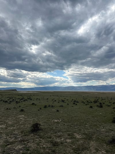 The image depicts a landscape scene. The sky is filled with dark, ominous clouds on the left side, contrasting with a brighter, sunlit area in the center, and lighter clouds on the right. A mountain range is visible in the middle distance, appearing as a narrow strip between the sky and the foreground. The foreground consists of a field, primarily green with patches of brown, indicating possibly dry or dormant vegetation. No people, recognizable subjects, actions, or interactions are discernible in the image. The location, Khuzhir, Russia, is not directly confirmed by the image content but is indicated as a known detail.