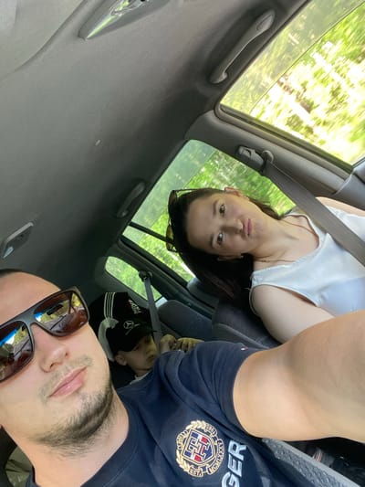 A selfie is taken from the perspective of the driver of a vehicle traveling through a wooded area. In the foreground, the driver, a man wearing sunglasses and a dark t-shirt with a logo, is partially visible. To his right, in the back seat, a woman wearing a white top makes a playful facial expression. A young boy, wearing a black baseball cap with a Mercedes-Benz logo, is also seated in the back. The car has a sunroof open, revealing the green trees and foliage blurred by motion outside. The interior of the car appears to be a typical passenger vehicle. The location is identified as Burdakovka, Russia.