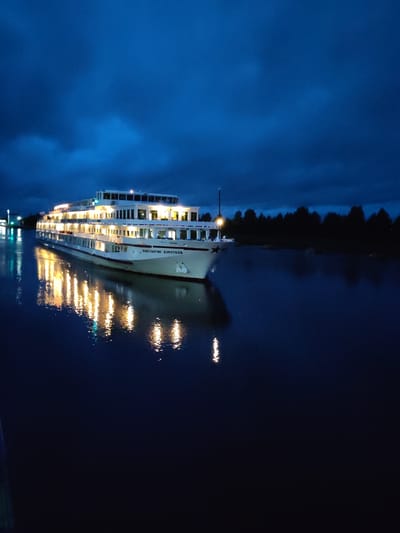 A large white river cruise ship named "KONSTANTIN KOROTKOV" is docked on a dark blue body of water, likely a river, under a deep blue, cloudy night sky. The ship is brightly illuminated from within, with numerous windows glowing warm yellow. Its reflection is clearly visible on the still water, creating a mirrored effect of the lights. The ship has multiple decks and a distinctive profile with a star emblem on its bow. A dark, silhouetted treeline forms the background on the right side of the image, suggesting a natural landscape. On the left side, a distant structure with lights is visible, possibly a dock or another building. The overall atmosphere is calm and serene, despite the dark sky. The location is identified as Devyatiny, Russia. There are no people visible in the image.