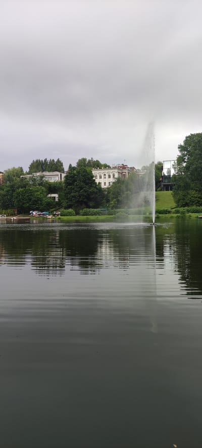 The image captures a tranquil scene of a lake with a tall fountain spewing water high into the air. The lake's surface is rippled, reflecting the overcast sky and the lush green trees that line the shore. Behind the trees, several multi-story buildings are visible, including a large white building with intricate architectural details and a more modern structure with glass walls. On the left side of the image, a small dock is visible, with several small boats and kayaks neatly arranged on it. The overall atmosphere is peaceful and serene, suggesting a well-maintained park or residential area. The location is specified as Angelovo, Russia.