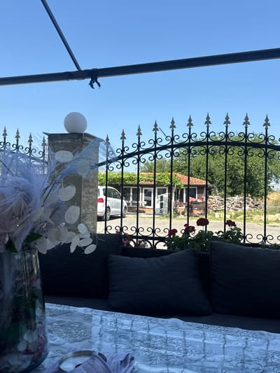 In Slanchevo, Bulgaria, an outdoor seating area is depicted with a black metal fence in the foreground. Behind the fence, a white minivan is parked in front of a small, rustic building with a red tiled roof. The scene is bathed in natural daylight under a clear blue sky. The seating area is furnished with dark-colored cushions on a bench, and a glass vase containing a floral arrangement is visible on a table covered with a patterned cloth.