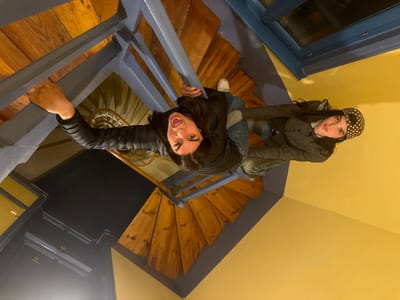 This image, rotated 90 degrees clockwise, depicts two individuals inside a building in Roanne, France, positioned on a multi-level spiral staircase. The staircase features wooden treads and blue-painted railings.

The subject on the left, with long dark hair, wears a black puffer jacket and blue jeans. They are shown gripping a blue handrail with their left hand, their mouth open wide and eyes wide, facing the camera. Below them, a dark doorway is visible on a landing, alongside the central dark void of the stairwell.

The subject on the right, also with long dark hair, wears a dark grey jacket and blue jeans, with a dark head covering adorned with white polka dots. They hold a blue handrail with their left hand, with their right foot, wearing a white shoe, visible on a wooden step. This individual maintains a neutral expression, looking towards the camera.

The background includes yellow-painted walls and a blue-framed window positioned in the upper right portion of the image.