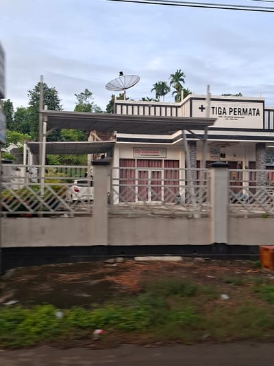 The image shows a building with "TIGA PERMATA" written on it in large letters, located in the city of Keude Lueng Putu, Indonesia. A satellite dish is visible on the roof of the building. In front of the building, there is a white fence and a white car parked under a canopy. The sky is cloudy, and there are trees in the background. The lower part of the image shows a muddy area with some debris. The overall scene appears to be a commercial establishment, possibly a clinic or a business, captured from a slightly elevated viewpoint, suggesting the photographer was in a vehicle.