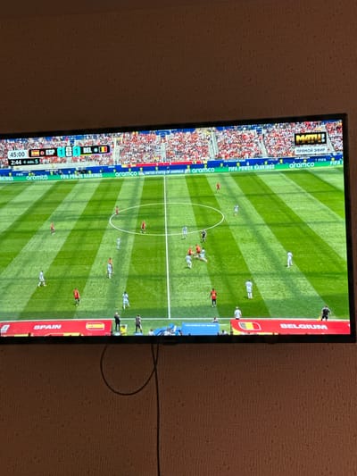 A television screen displays a soccer match between Spain and Belgium. The score is tied 1-1 with 45:00 minutes played plus 2:44 of added time. The game appears to be a live broadcast from a stadium with a crowd in the background. The screen also shows sponsorship logos such as "aramco," "FIFA," and "MATH!". The television is mounted on a wall with a textured beige wallpaper. A black cable hangs down from the bottom of the television. The image is taken from a slightly elevated angle, looking down at the television.
