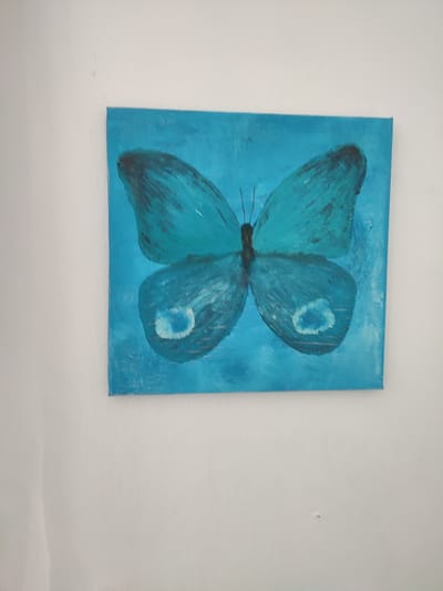 A painting of a blue butterfly is displayed on a white wall. The butterfly is the central focus of the image, rendered in various shades of blue with darker accents on its wings. It is depicted with its wings spread open. The background of the painting is a solid, textured blue. The white wall appears to be a typical interior wall, possibly in a home or gallery setting. There is no indication of the specific location in Денисово, Russia, within the provided image.