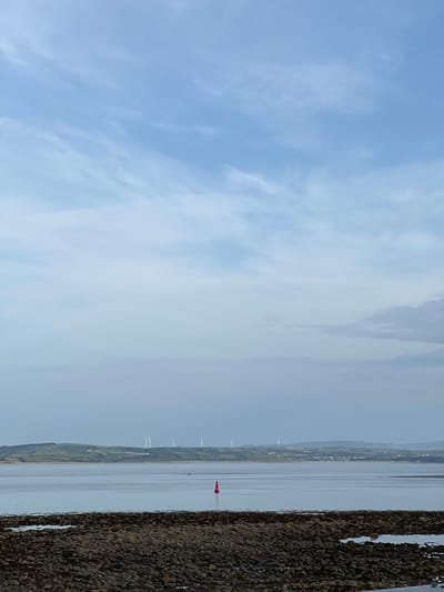 This is a landscape photograph taken from a rocky shoreline, looking out over a calm body of water towards a distant coastline. The sky is a pale blue with wispy white clouds.

In the water, a bright red buoy stands out against the muted tones of the sea and distant land. Further inland, a line of green hills is visible, dotted with what appear to be houses and wind turbines. The presence of wind turbines suggests an open, likely coastal area.

The foreground is dominated by a textured surface of dark, rounded stones and pebbles, suggesting a beach or harbor wall. The water is still, reflecting the sky and the distant landscape.

The lighting suggests it is daytime, possibly late morning or early afternoon, with a clear but not overly bright sun. The overall atmosphere is serene and tranquil. There is no visible text in the image. The scene evokes a sense of peaceful observation of a coastal environment.