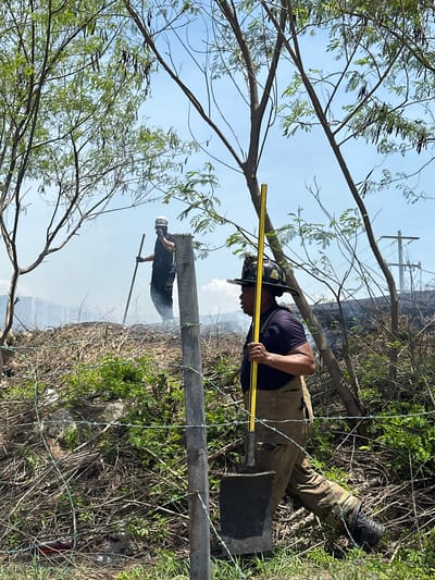 Two firefighters are visible in the image, working in a dry, brush-filled area under a bright blue sky. The firefighter in the foreground, closer to the camera, wears a dark shirt, tan overalls, a firefighter's helmet, and black boots. He is holding a yellow shovel with a black blade, angled downwards. His body is partially obscured by a barbed wire fence and some tree branches.

In the background, another firefighter is seen standing on a pile of debris, holding a long pole or tool. He is wearing a dark outfit and a white helmet with goggles. Smoke is rising behind him, suggesting a fire or smoldering debris. The environment is overgrown with dry brush and branches, with trees interspersed throughout. In the distance, a utility pole with power lines is visible against the sky, and a large building or structure can be seen through the haze. The scene appears to be located outdoors, in an area with vegetation and some man-made infrastructure. The overall impression is one of an active firefighting effort in a rural or semi-rural setting.