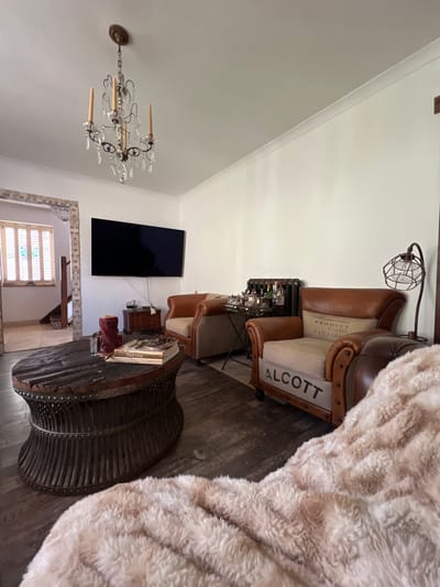 The media file shows a dimly lit living room with a masculine, industrial aesthetic. The scene is dominated by a large, plush, cream-colored faux fur blanket draped over a leather armchair. The armchair itself is a prominent object, featuring brown leather upholstery and some text on a cream-colored cushion that reads "PRODUCT BY CARGO" and "CAUTION". To the left of the armchair, a floor lamp with a caged bulb fixture is visible.
In the background, a flat-screen television is mounted on a white wall. Below the television, a distressed wooden coffee table with a metal rim and rivets sits on a dark wood floor. On the coffee table, a stack of books is visible, one of which has "Varmint" printed on its spine. A red decorative object is also placed on the table.
To the left of the coffee table, a clear crystal chandelier hangs from the ceiling, casting a soft light. The overall impression is one of a cozy, yet stylish, living space with a focus on texture and industrial-inspired decor. The lighting suggests it is either evening or the room has ambient lighting. No people are visible in the media file. There are no specific weather cues or strong location cues other than the provided context of Brighton, United Kingdom.