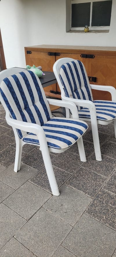 The image depicts a scene likely in an outdoor area, possibly a patio or garden, given the paving stones on the ground. Two white plastic chairs with blue and white striped cushions are the primary focus. To the left of the chairs, a dark table is visible, with a light green plush toy resting on it. Behind the chairs and table, a wooden chest or cabinet with dark hardware is positioned against a white wall. A window with frosted glass panes is present in the upper right portion of the image, set into the white wall. A small yellow bird with blue markings is perched on the windowsill. The overall impression is one of an outdoor seating area, possibly in a residential setting in Dietzenbach, Germany.