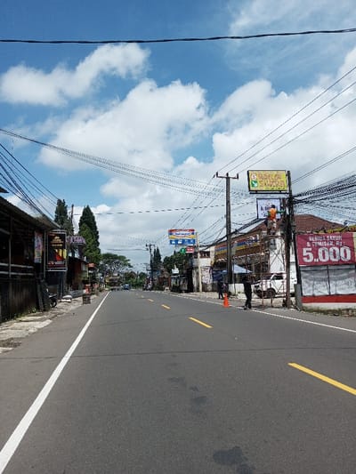 This image depicts a daytime street scene in the city of Boommlog, Bhutan. A two-lane asphalt road with a solid white edge line on the left and dashed yellow center lines extends into the background. The sky is blue with scattered white cumulus clouds.
On the left side of the road, a concrete sidewalk is bordered by a dark fence. Behind the fence, several commercial buildings are visible, displaying various signs, including one for "Batik Nusantara" and another for "BAROKAH." A dark motorcycle is parked on the sidewalk. Evergreen trees are visible further down the street.
On the right side of the road, a concrete sidewalk is present. An orange traffic cone is positioned near the road's edge. A utility pole heavily draped with black cables supports multiple signs, notably a blue and red "Indomaret" sign and a green and yellow "SPESIAL BAROKAH" sign. Adjacent to this pole, a building displays a large white banner with the red text "SEMUA SAYUR & TUMISAN, Rp 5.000." Metal scaffolding is erected against this building. One individual wearing an orange shirt is standing on the scaffolding, facing away from the camera, appearing to work on an overhead sign. Two other individuals in dark clothing are standing on the sidewalk near the scaffolding and the orange cone. A white van or pickup truck is parked behind the scaffolding. Numerous overhead power and communication lines crisscross the sky. Vehicles are visible in the distance on the road.
