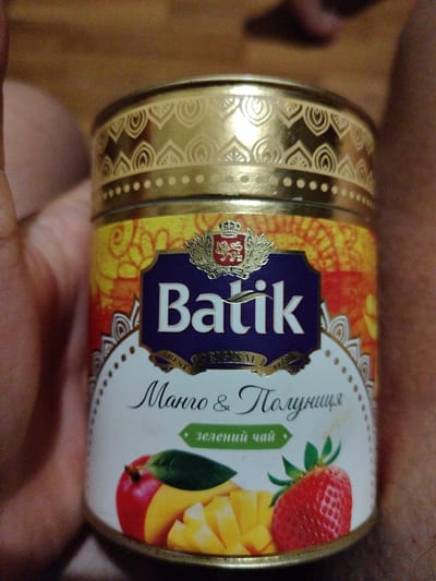 A close-up, eye-level shot shows a round tin of Batik brand tea held by a person. The tin features a gold, ornate patterned lid. The main label is white and blue, with the text "Batik" prominently displayed in white lettering on a blue background. Below this, in a script font, is "Mango & Клубника" (Mango & Strawberry) and then "зеленый чай" (green tea) in a green banner. Illustrations of mango slices, a strawberry, and what appears to be a red apple or another fruit are present on the lower part of the label. The background is a blurred wooden surface and parts of the person holding the tin. There is no discernible information about the location in Okhtyrka, Ukraine.