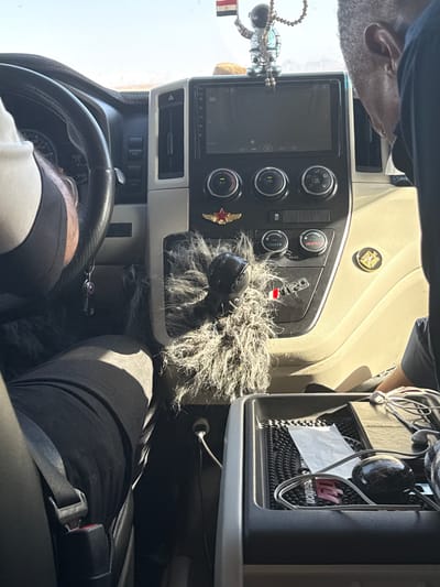 The interior of a vehicle, possibly a truck, is shown. The driver, a person with gray hair and tan skin, is partially visible in the upper left corner of the frame. The dashboard, steering wheel, and center console are prominent. A furry cover surrounds the gear shift. On the dashboard, there are control knobs and a digital screen. A red star emblem is pinned to the console near the gear shift. A tissue box is visible to the right of the console. The driver is wearing a black shirt and has a seatbelt fastened across their lap. The image was taken inside the vehicle; location information is not discernible from the image content.