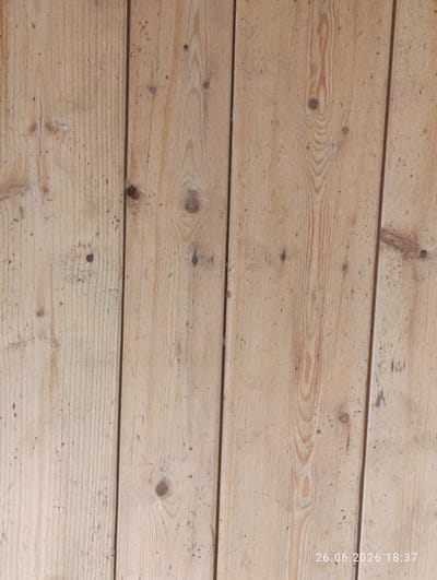 This is a close-up, eye-level photograph of a wooden surface composed of vertical planks. The wood appears to be unfinished pine, exhibiting a light brown color with prominent grain patterns and occasional knots. Small dark speckles, likely dirt or imperfections in the wood, are scattered across the surface. The planks are joined together, forming a textured and slightly uneven background.
The image is taken indoors, as indicated by the artificial lighting which casts a uniform, soft glow on the wood. There are no people or distinct objects present, focusing solely on the texture and details of the wood. The scene is static and devoid of any specific activity.
In the bottom right corner of the image, a timestamp is visible, reading "26.06.2026 18:37". This suggests the photograph was taken in the early evening of June 26, 2026. The location context provided is Novovolynsk, Ukraine. The overall impression is one of a simple, rustic, and functional surface.