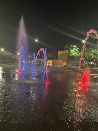 The image captures a night scene at the Lagos water fountain, a prominent attraction in the city of Lagos, Portugal. The fountain is illuminated with vibrant red and blue lights, creating a dynamic visual display as water shoots upwards in various patterns. The water reflects the colorful lights onto the dark, rippling surface of the fountain's basin, adding to the magical ambiance. In the background, a historic stone wall with crenellations suggests the presence of a castle or ancient fortification, a common feature in Lagos. Lush green trees frame the scene, with palm trees also visible, hinting at the Mediterranean climate. A bright moon hangs in the dark night sky, its soft glow contributing to the overall atmosphere. A few individuals are seen in the background, appearing to be enjoying the spectacle, some seated on a bench. The lighting design of the fountain, with its contrasting colors and the upward spray of water, makes it a captivating centerpiece of the urban landscape.