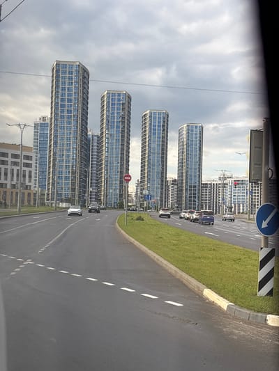 The image depicts a wide, multi-lane asphalt road with clear white lane markings, including a dashed line in the center and solid white lines on the right edge. The road curves gently to the right. On the right side of the road, there is a well-maintained grassy area with a concrete curb and a striped black and white pole with a circular blue sign indicating a downward arrow. Beyond the grassy area, several modern, tall, octagonal apartment buildings with many blue-tinted windows are visible under a cloudy sky. In the distance, more buildings of varying heights, some with white facades and grid-like window patterns, fill the skyline. A traffic light is visible on the right. Several cars are driving on the road, including a white sedan, a dark SUV, a blue minivan, and other vehicles. The overall impression is a contemporary urban environment. Based on the architectural style and the presence of tall apartment buildings in a suburban setting, this scene is likely in the vicinity of the Сеніцкі сельскі Савет in Belarus, a region known for new residential developments.